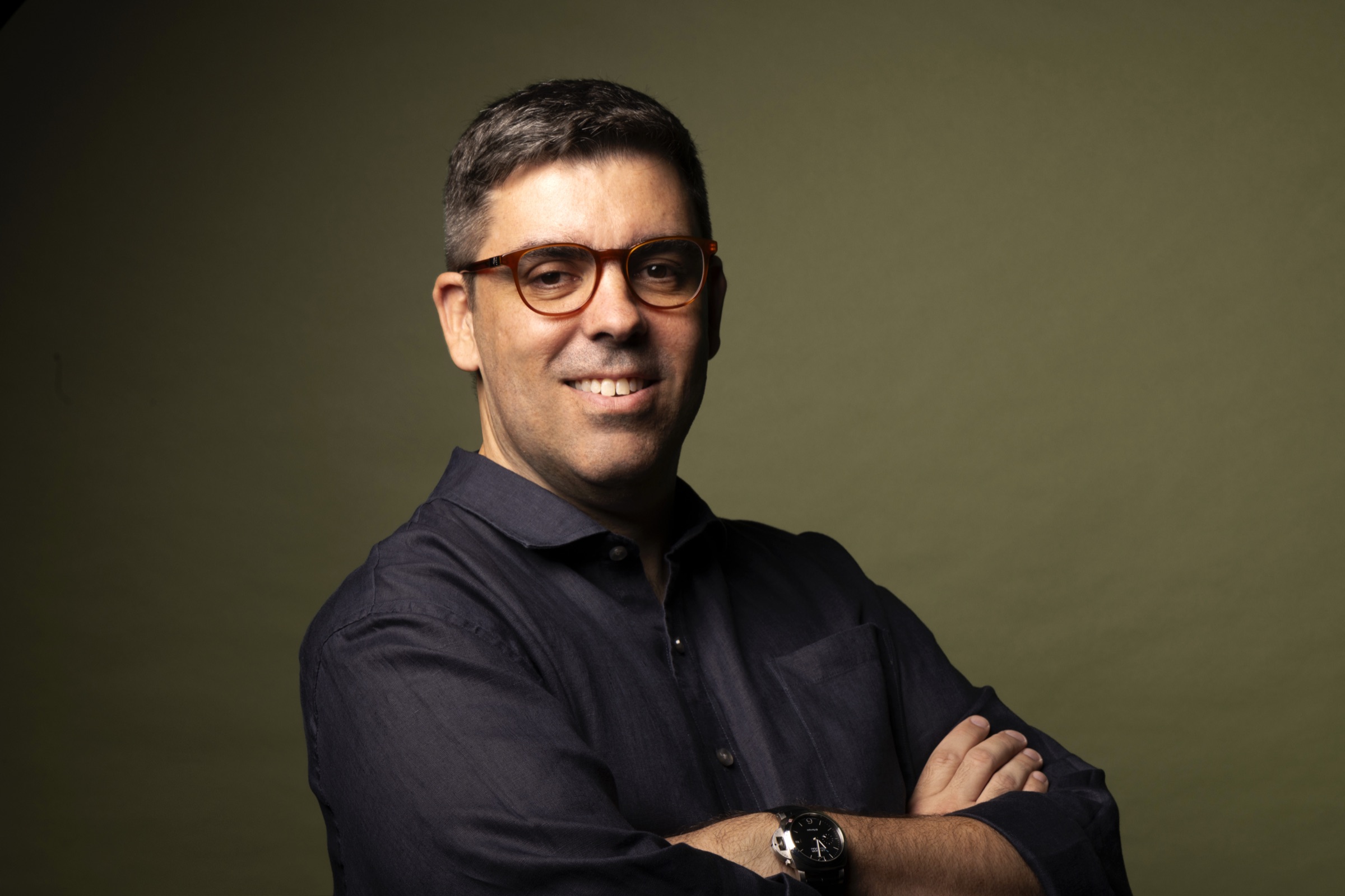 Eduardo Cruz sorrindo, de braços cruzados, em fundo verde-oliva
